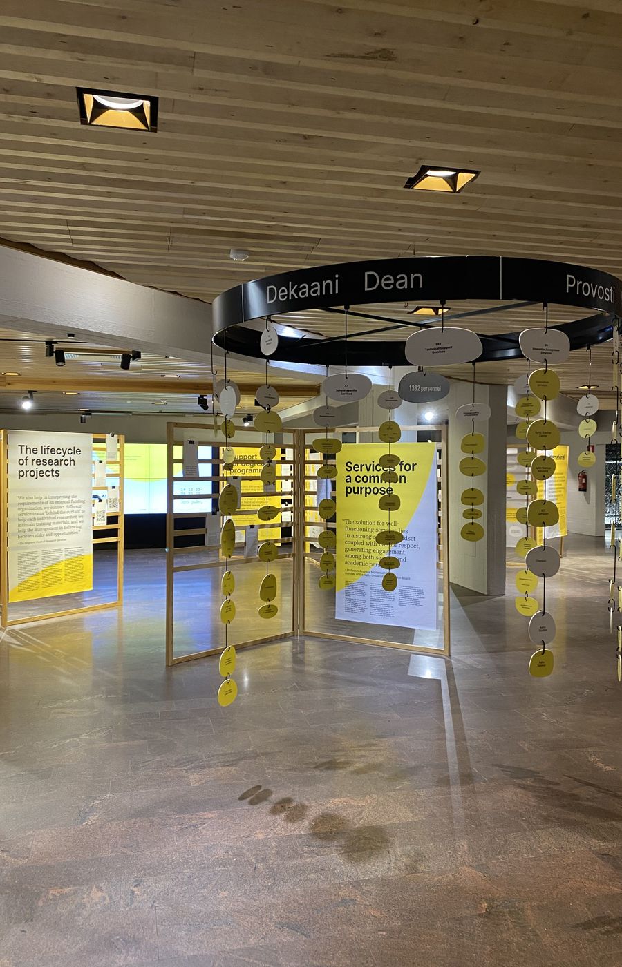 exhibition installation view, large mobile hangs in right foreground, behind wooden stands withprinted yellow and gray fabrics