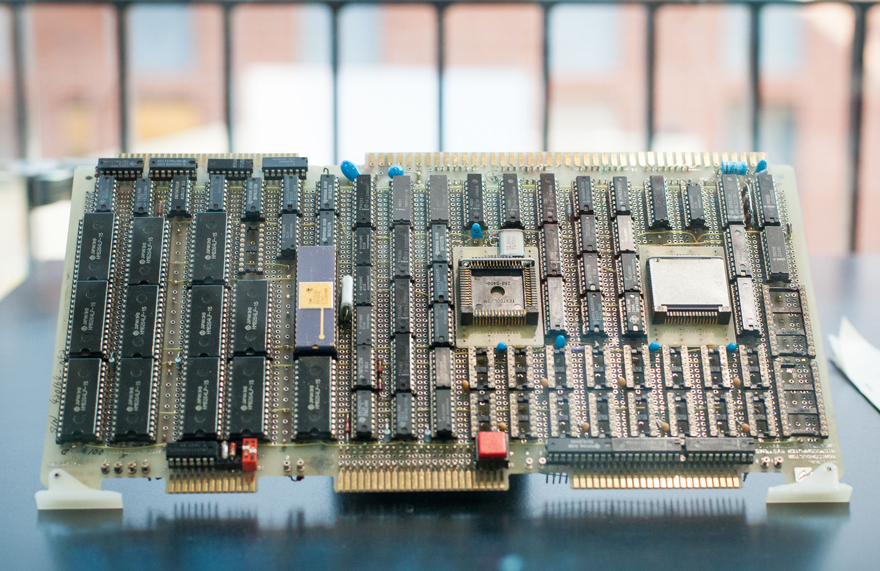 A computer control board is in the centre of the frame on a black base. the board is covered in metallic computer chips and behind it are railings out of focus. 