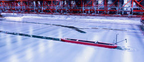 A red model ship breaks through the ice from left to right of the image middleground. In the background the red metal bridge is visible across the full width of the image. 
