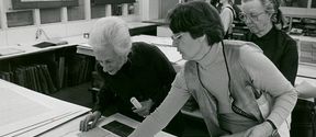 Two women on the right of the image lean over a table to inspect materials. Another women stands behind them on the far right. Three students stand together in the background. 