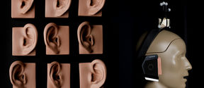 On the left are 9 plastic models of ears in varying flesh tones are laid on top of black fabric in a grid formation. On the right, A beige toned plastic model of a human head and shoulders is in profile view, facing to the right of the image. A pair of over-ear headphones rest on the model head 