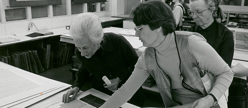Two women on the right of the image lean over a table to inspect materials. Another women stands behind them on the far right. Three students stand together in the background. 