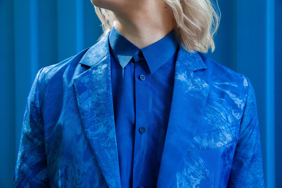 A blonde woman in a blue suit jack and blue shirt is centre frame. Behind her, a blue background. Only her neck and torso are visible in the image.
