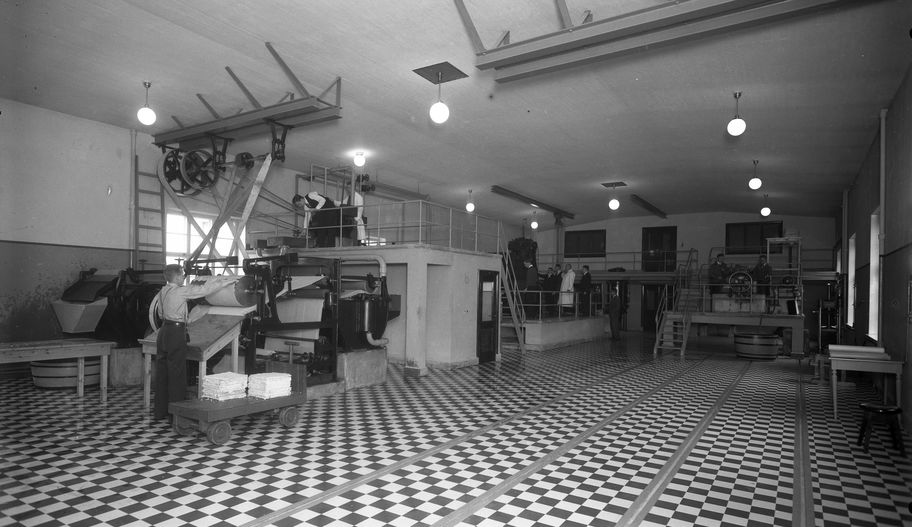 In a large laboratory with a black and white tiled floor, there are large machines. A man works a large machine on the left of the image which has a giant roller and a second level above to access the upper part of the machine. Pictured is Johannes Brax, a professor of paper technology, with students using different machines in the lab.