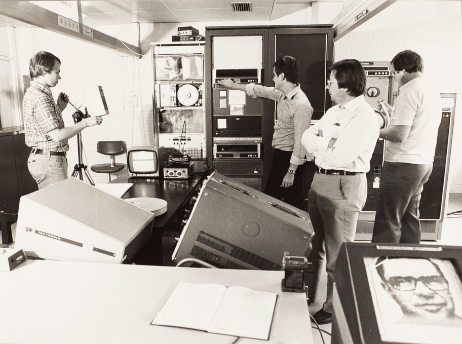 Teuvo Kohonen is pictured in the right middle ground of a sepia coloured photograph. Three other members in his research group stand in the background. There are various machines and instruments against the back wall of the room and also in the foreground. 