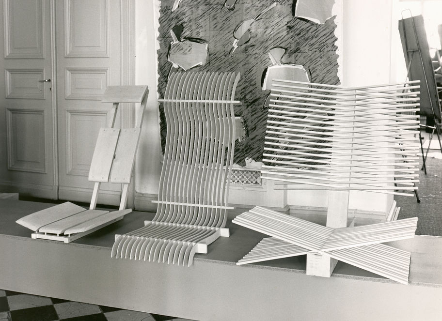A selection of three chair designs from the 1980s sit on a raised platform. Behind them a painting hangs on the wall. The back of a painting easel is visible in the right background of the image. 