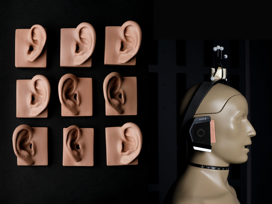 On the left are 9 plastic models of ears in varying flesh tones are laid on top of black fabric in a grid formation. On the right, A beige toned plastic model of a human head and shoulders is in profile view, facing to the right of the image. A pair of over-ear headphones rest on the model head 