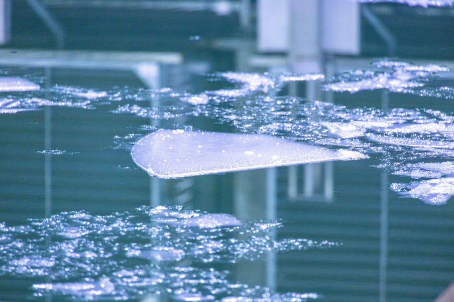 White cloudy ice starting to form on the surface of the water in the Ice Tank. In the centre middlegrounud of the images, a triangular plate fo ice is forming. In the foreground the ice is still crystalizing. The unfrozen water suraface is blue green in colour and reflects the ceiling above. 