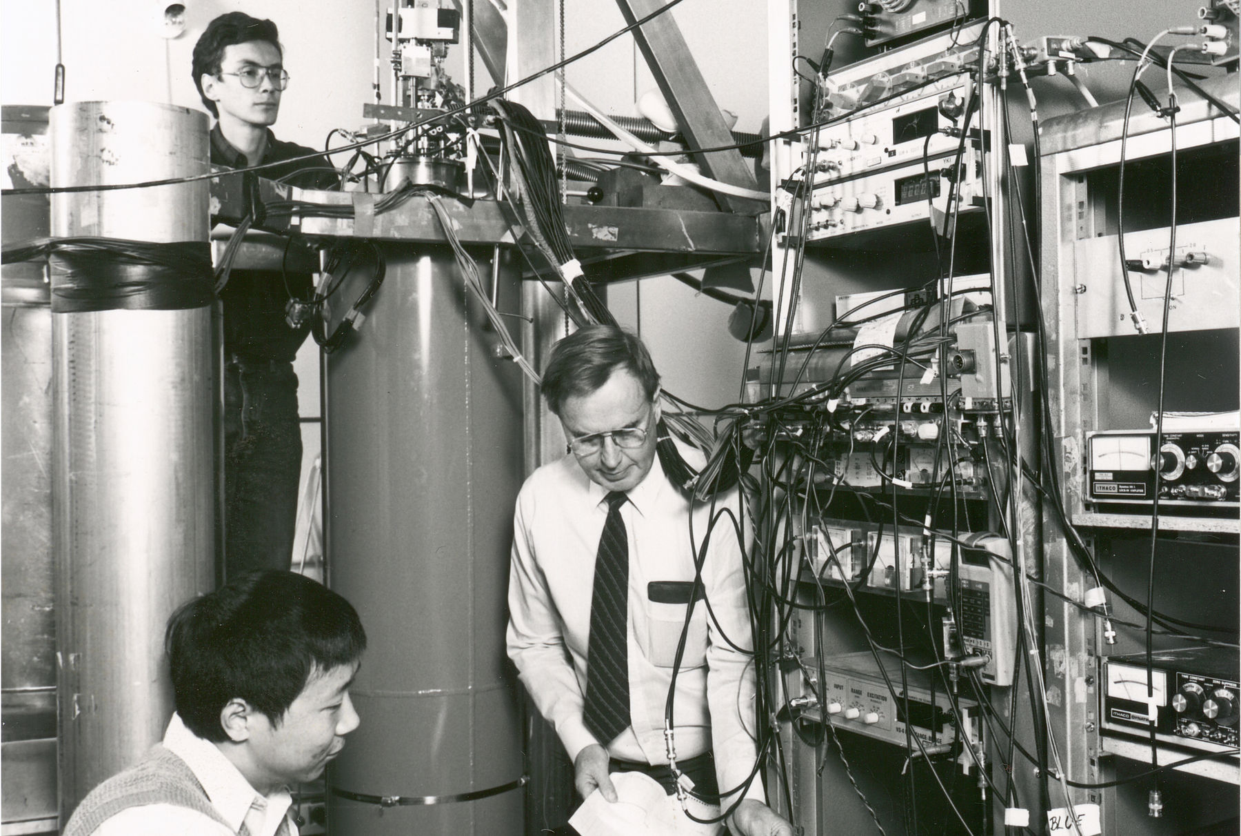 The picture shows three researchers in the Low Temperature Lab. Two men check the progress of the study on a machine. One stands behind a large cylinder.