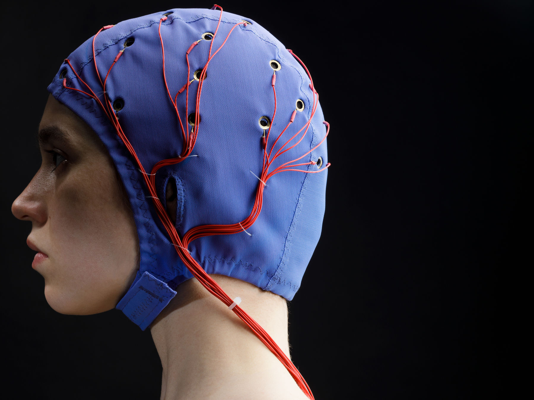 A woman in profile view facing left wears a cobalt blue EEG cap with red wires coming from circular metal eyelets on the surface. She is slightly left of centre. Her hair is not visible under the cap. The flash coming from the right illuminates the back of her head and leave her face in shadow. The red wires fall from outside her ear to behind her shoulder.