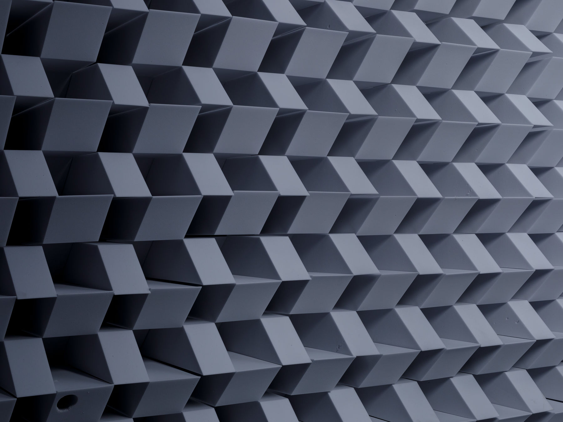 The full frame shows the surface of a soundproofing wall in an anechoic chamber. Gray pyramids of foam cover the surface creating a geometric pattern. The flash from the camera renders high contrast between the shapes, starting darker in the image background and gradually lightening as it comes to the foreground.